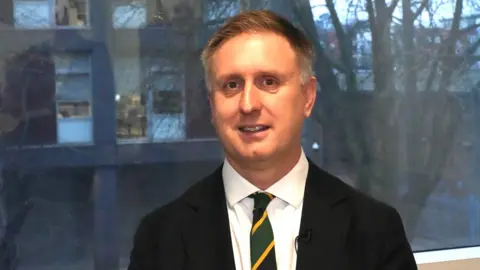 John Fairhall/BBC Ed Garratt smiles at the camera in front of a large window. He wears a black suit with a green and yellow tie. He has short grey hair.