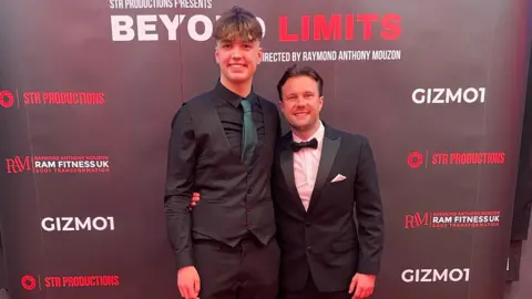 A tall young man wearing a black suit and a dark green tie and a slightly older man wearing a tuxedo. They're standing with their arms around each other and smiling at the camera, with a banner for their film behind them.