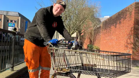 Henry Godfrey-Evans/BBC A man in a jumper and high-visibility trousers holding a trolley with gloved hands next to a river bank.