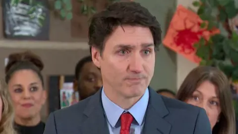 Justin Trudeau wells up during one of his last press conferences as leader of Canada. He is wearing a steel blue suit, red tie and blue shirt. Four people are visible standing behind him.