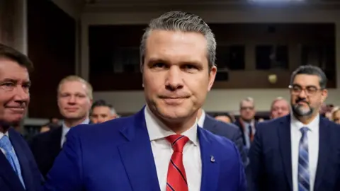 Pete Hegseth stares into the camera with several men in suits behind him.