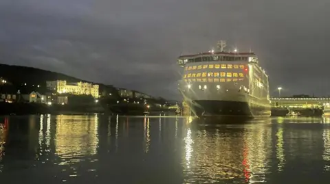 BBC The Manxman, which is a large red, white, and black ferry, sitting in low water in Douglas Harbour at night. The lights from the ship, the harbourside buildings are reflecting on the water around it.