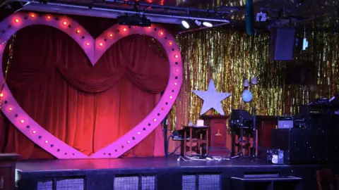 A stage with a large pink heart with gems around it against red curtains, next to gold foil curtains with a white star propped up against the wall.  