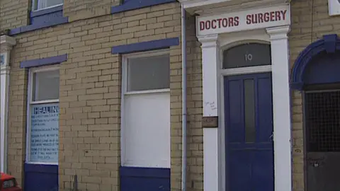 The front of a brick-built GP practice. The building has a blue front door with a sign above it reading Doctors Surgery.