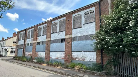 A side photo of an empty building. The sky is blue and the building is red brick with five pillars. In between each pillar is bordered up windows with blue and grey boards.