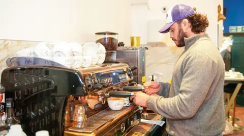 Seth Conway Media A man concentrating making coffees using a coffee machine. He's wear a grey jumper with a purple hat. The coffee machine has lots of cups above it with coffee beans stored alongside. 