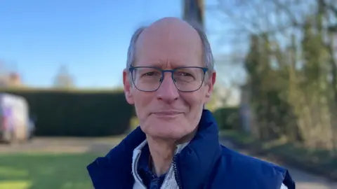 Luke Deal/BBC Churchwarden Richard Chatham smiles at the camera. He is largely bald with some grey hair around the side of his head. He wears a blue gilet with a white jumper underneath.