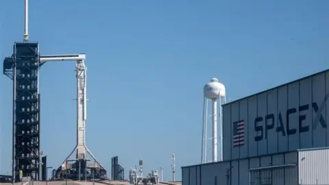 The rocket ahead of launch. Sitting in its stand to prepare to launch, a large building is visible in the near ground with SpaceX written on its side 