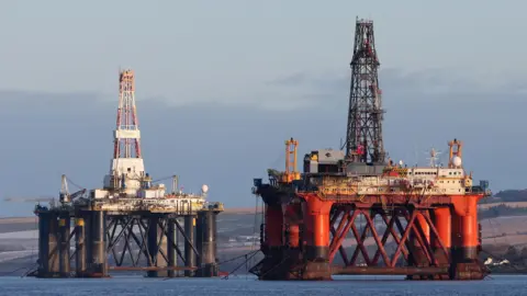 PA Media Two oil platforms in the North Sea. The one in the foreground is red and has a white cabin with a black tower on top. In the background, the platform is black with a white cabin and white and red tower.