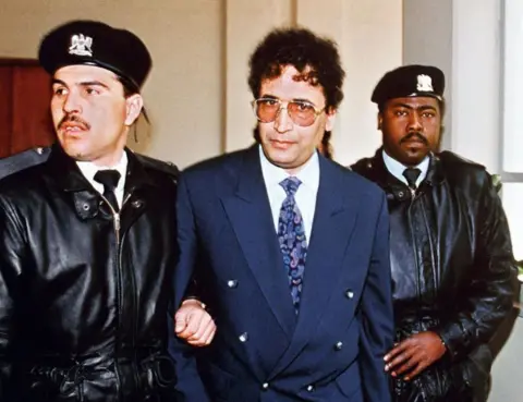 Getty Images Abdelbasset al-Megrahi is escorted by two security officers in Tripoli in 1992. He is wearing a blue suit jacket, a white shirt, a patterned blue and purple tie and has large glasses. The officers are dressed in black coats and black berets.