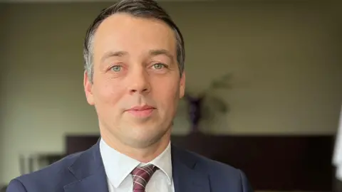 Filip Hermann is sitting in a hotel conference room. He is wearing a navy suit and red, checked tie and is looking at the camera.