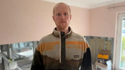 BBC Liam Reilly who has short ginger hair and a beard and is wearing an orange and brown Columbia fleece. He is stood in a room with renovation works going on behind him.