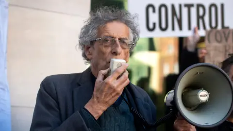 Victoria Jones/PA Wire A man in his 70s with white hair and wearing a grey jacket speaks into a megaphone at an outdoor protest. 