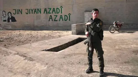 A man in army uniform stands in front of a building holding a gun. 