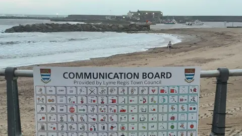 Lyme Regis Town Council A large board attached to the railings on the beach. It has 144 squares, each containing a commonly used word or number with an illustration. Behind the sign is the sandy beach and sea with the Cobb harbour wall in the background.