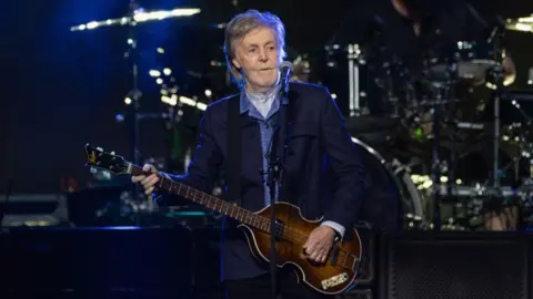 Paul McCartney wearing a blue jacket and blue shirt performing on stage with his left-handed guitar at the O2 Arena.