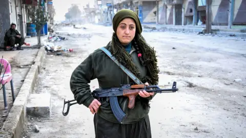 Getty Images A woman holding a gun stands in a destroyed street in Kobane. There are wrecked buildings behind her.   