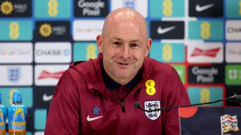 Getty Images Lee Carsley in a maroon red England jacket, sitting at a table with a microphone in front of him. He is smiling. He is bald and has a blue flower pinned on his jacket