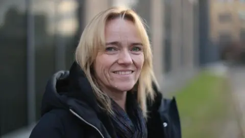 BBC A woman with shoulder-length blonde hair smiles at the camera. She is wearing a black jacket and appears to be standing outside a building.