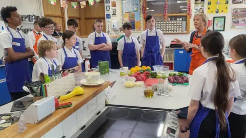 A group of students in white shirts and blue aprons stand in a kitchen classroom, listening to Kate, who is wearing an orange apron, speak. On the table there is a number of cutting boards with peppers and onions on them. 