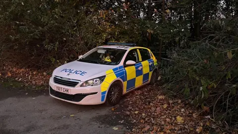 A police car parked at the entrance to a field surrounded by a hedgerow