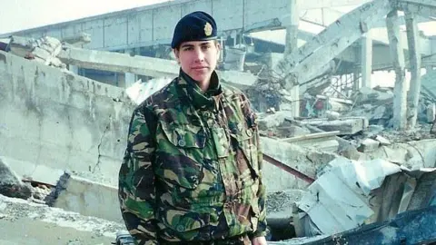 Gemma Morgan Gemma Morgan wearing camouflage uniform and a black military cap. She is standing amongst rubble and a destroyed building while on active duty. She is looking at the camera with a blank expression