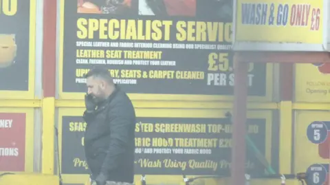 National Crime Agency Khdir in a black coat and is on the phone. He is in a car wash where there are yellow signs offering differnt car wash services.