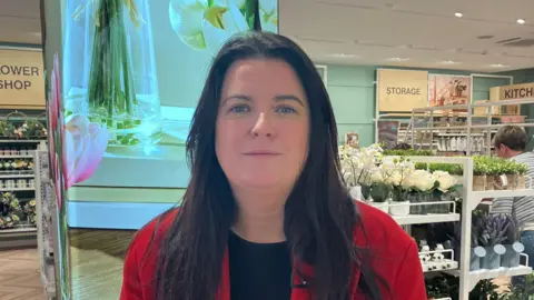 Martina Connolly. She has long dark hair, wearing a red blazer with a black top underneath. Primark home decor can be seen in the background.