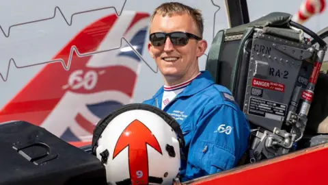 Sgt Harry Kay wearing a blue overall and sunglasses, sitting in a Hawk jet holding a white helmet. He is smiling and has short fair hair.